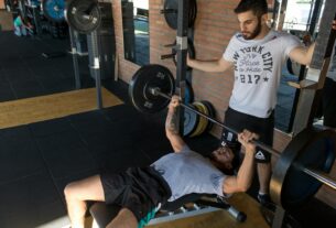 A strong man lifts weights on a bench while a trainer assists in a modern gym setup.